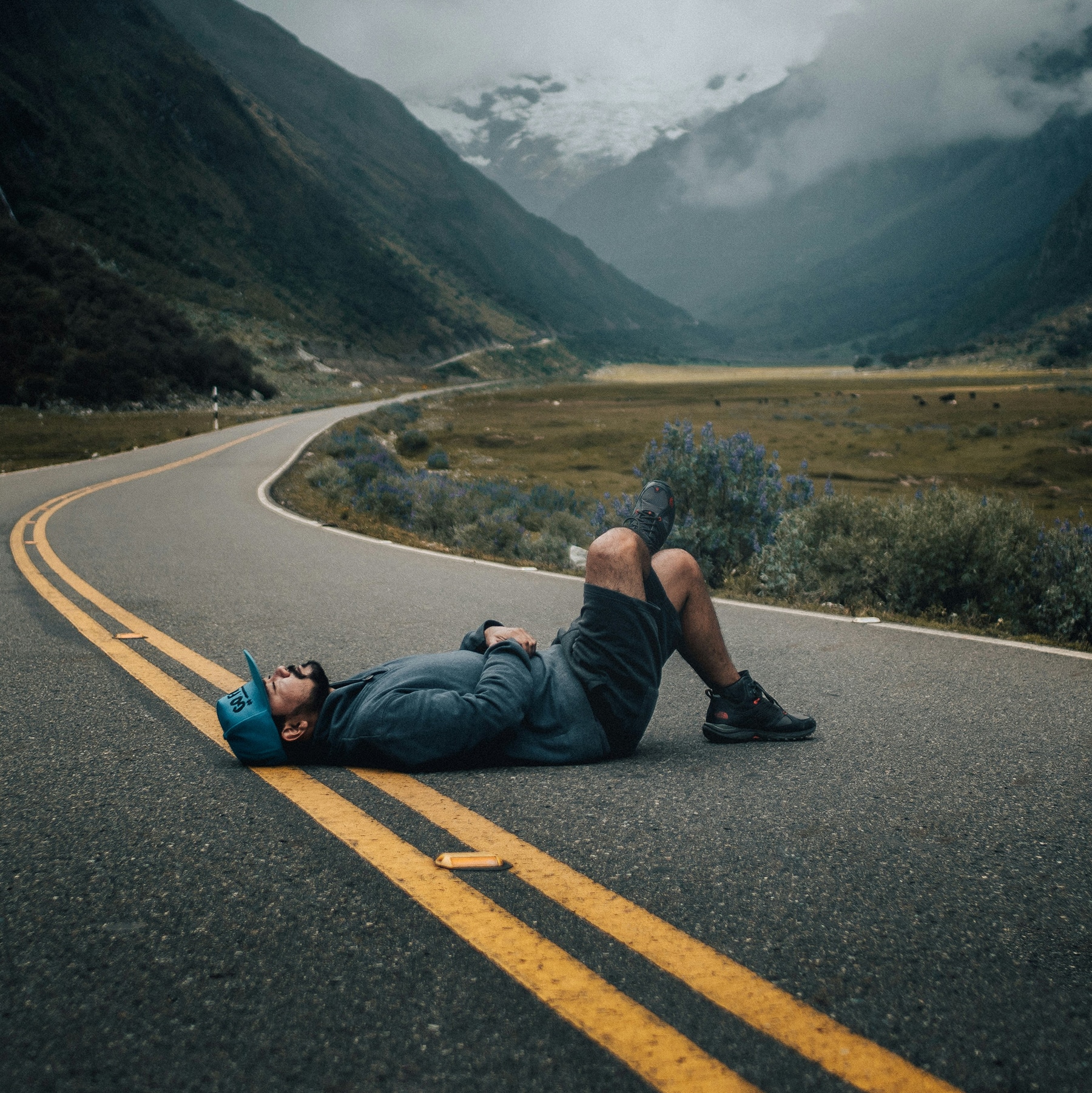 Midlife man lying on his back on a mountain road, resting mid-journey, with a misty green valley and mountains behind him