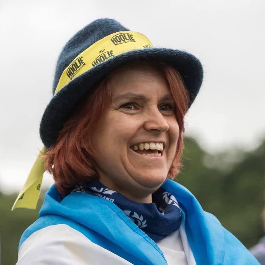 Portrait of Martha smiling during a music festival. She is wearing a hat and is wrapped in a Scottish flag